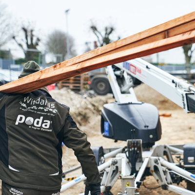 Montør bærer lange treplanker på en byggeplass med lift og materialer.