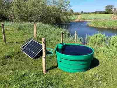 Soldrevet feltpumpe med solcellepanel og vanntank ved en bekk på en eng.