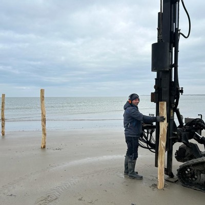 Montør setter opp trestolper med maskin på en strand ved kysten.