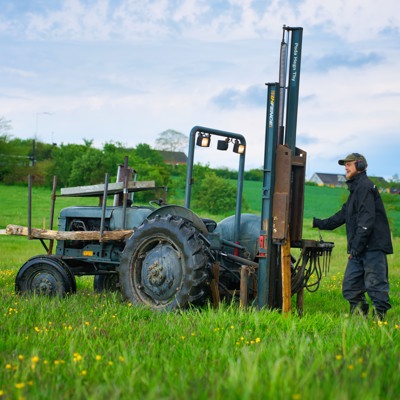 Montør arbeider med pålehammer på traktor ved oppsetting av gjerdestolper i et grønt jordbruksområde.