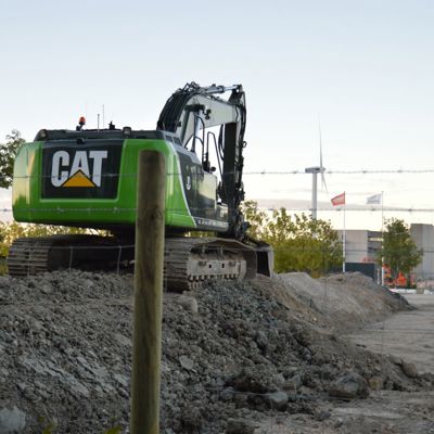 På en byggeplass står en grønn CAT-maskin parkert. For å øke sikkerheten på byggeplassen er det satt opp et byggeplassgjerde