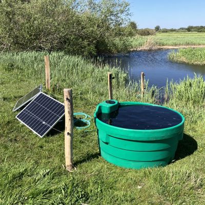 Soldrevet feltpumpe med solcellepanel og vanntank ved en bekk på en eng.