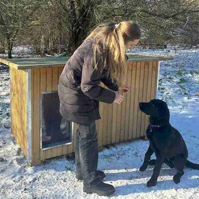 Kvinne gir en svart hund en godbit foran et hundehus i et vinterlandskap med snø på bakken.