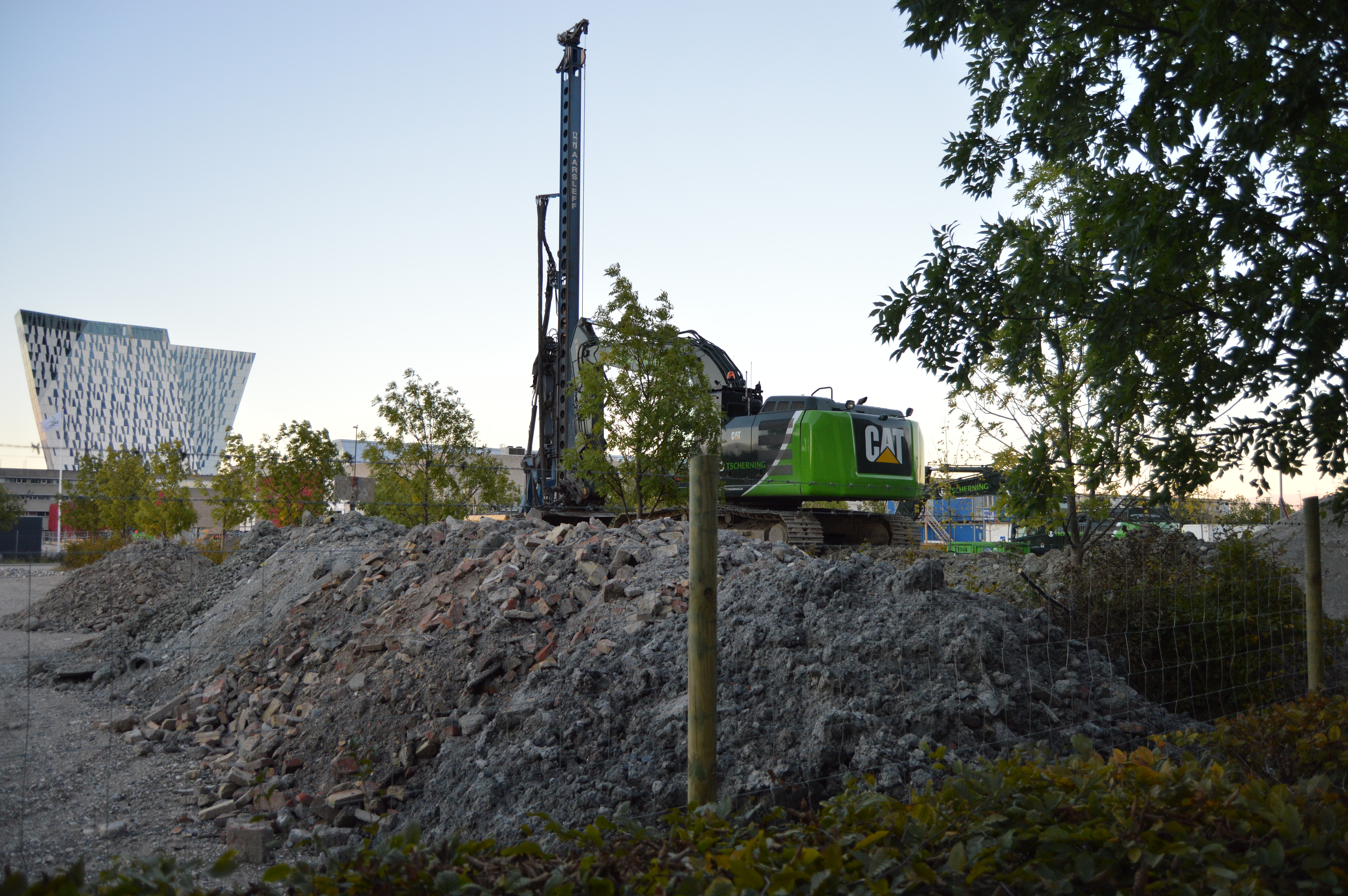En grønn CAT-maskin står parkert på toppen av en grushaug. Maskinen er sikret med et byggeplassgjerde.