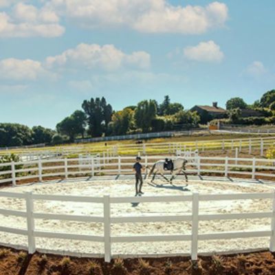 En hestetrener fører en hvit hest rundt i en roundpen. Roundpen består av et hvitt hestegjerde laget av hvit plast.