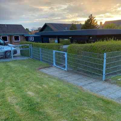 Panelgjerde med en port montert langs en sti som skiller en plen fra en hekk. I bakgrunnen sees en carport, flere hus og en solnedgang.